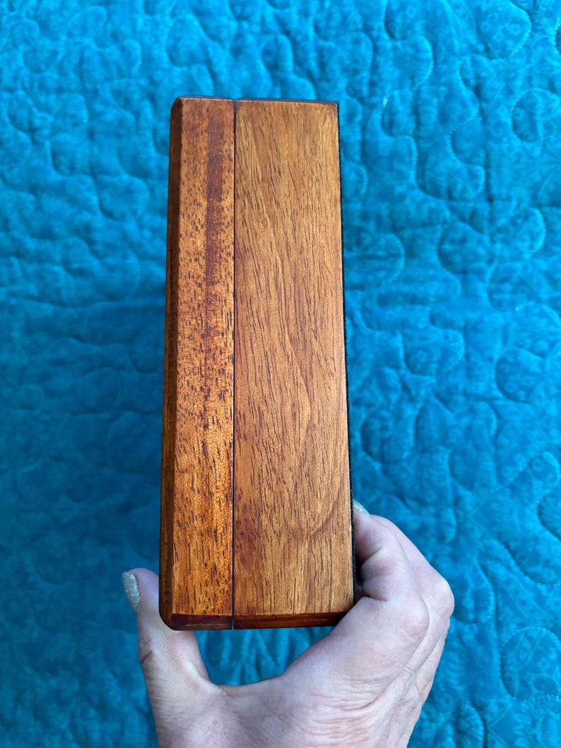 K&ouml;nnte beinhalten: Eine rechteckige Holzkiste mit Scharnierdeckel, aus rotbraunem Holz gefertigt. Die Kiste wird in einer Hand vor einem blauen, gesteppten Hintergrund gehalten. Die Holzmaserung ist sichtbar und verleiht dem Design Textur.