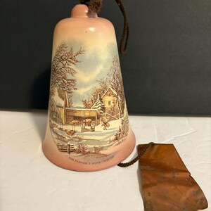 May include: A ceramic bell with a winter scene illustration. The bell is pink and features a brown rope for hanging and a copper tag. The illustration depicts a farm with a barn, trees, and snow, with the text "THE FARMER'S HOME - WINTER".