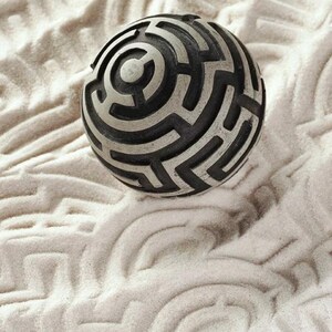 May include: A spherical object featuring a raised, intricate black and silver maze pattern across its surface. The design consists of interconnected paths and channels that form a complex labyrinthine structure, creating a high-contrast visual effect.