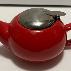 May include: A red ceramic teapot with a stainless steel lid and a handle. The lid has a small metal latch.