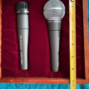 K&ouml;nnte beinhalten: Zwei graue Shure-Mikrofone in einer Holzkiste mit rotem Samtinnenraum. Die Mikrofone sind mit "75th Anniversary Limited Edition" beschriftet. Die Box ist etwa 20 cm lang, wie ein Ma&szlig;band zeigt.