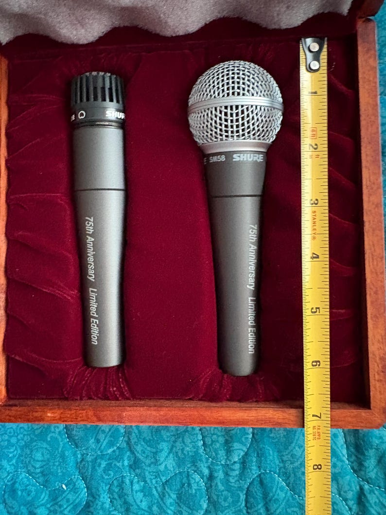 K&ouml;nnte beinhalten: Zwei graue Shure-Mikrofone in einer Holzkiste mit rotem Samtinnenraum. Die Mikrofone sind mit "75th Anniversary Limited Edition" beschriftet. Die Box ist etwa 20 cm lang, wie ein Ma&szlig;band zeigt.