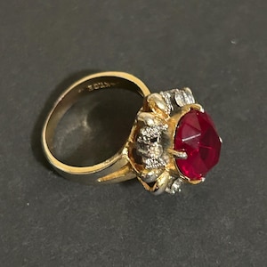 May include: A gold ring with a large red gemstone surrounded by smaller clear stones.