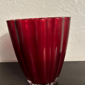 May include: A red glass vase with a fluted design. The vase is tall and slender, with a rounded base.