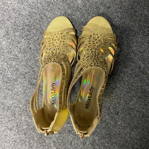 May include: A pair of gold sparkly heeled sandals with a decorative design. The sandals have a strap across the toes and an ankle strap. The brand name "Mila Lady" is visible on the inside of the sandals.