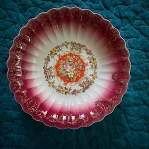 May include: A decorative porcelain bowl with a scalloped rim. The bowl features a white interior with a floral design in the center, surrounded by a red and gold border. The exterior is a gradient of red and white.