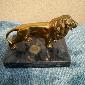 May include: A golden lion figurine on a dark gray marble base. The lion is in a walking pose with detailed mane and body. A small, circular emblem is on the base. The figurine is a decorative item.