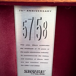 K&ouml;nnte beinhalten: Eine silberne Plakette zum 75-j&auml;hrigen Jubil&auml;um von Shure mit den Zahlen "57/58". Die Plakette befindet sich vor einem tiefroten Samtinterieur, mit Text, der die Geschichte des Unternehmens und die klassischen Modelle SM57 und SM58 beschreibt. Das Shure-Logo befindet sich unten.