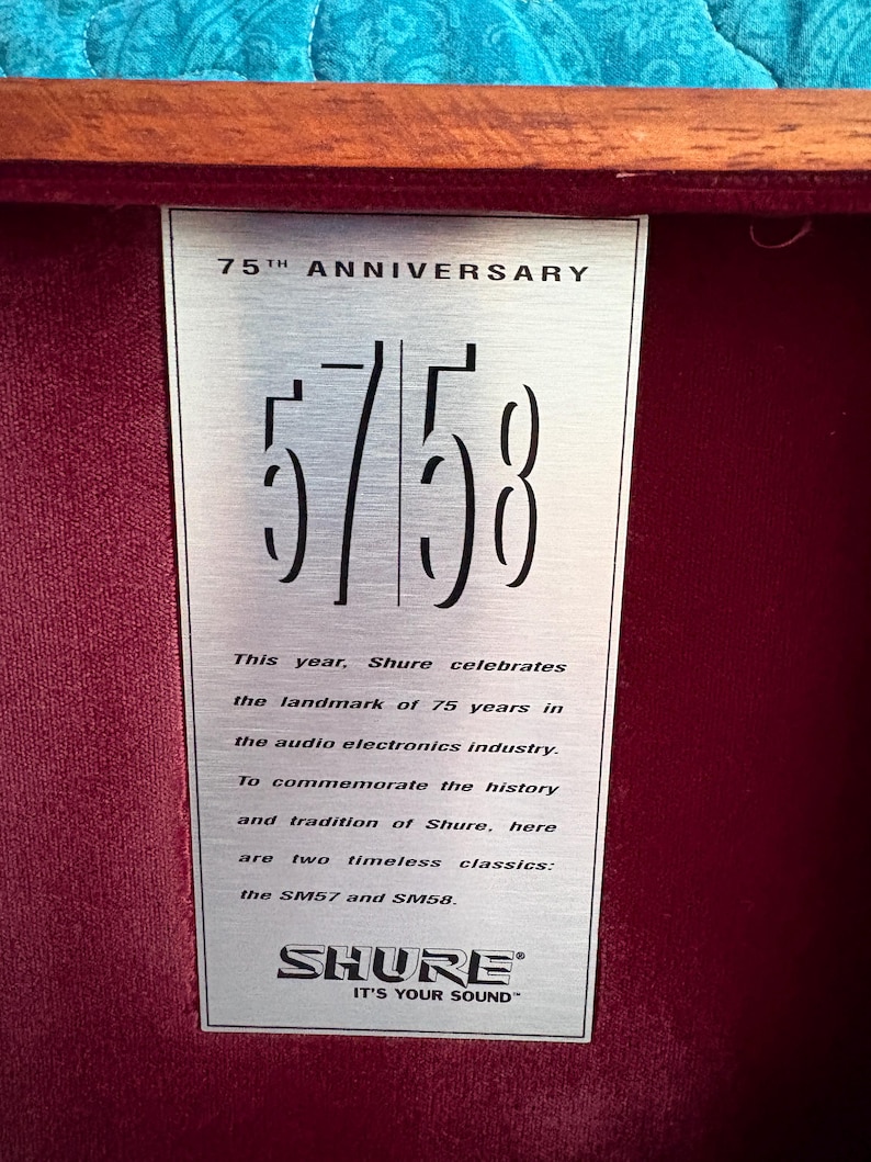 K&ouml;nnte beinhalten: Eine silberne Plakette zum 75-j&auml;hrigen Jubil&auml;um von Shure mit den Zahlen "57/58". Die Plakette befindet sich vor einem tiefroten Samtinterieur, mit Text, der die Geschichte des Unternehmens und die klassischen Modelle SM57 und SM58 beschreibt. Das Shure-Logo befindet sich unten.