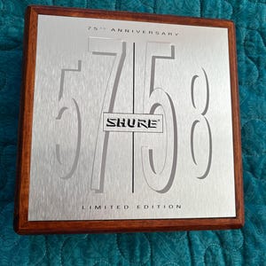 K&ouml;nnte beinhalten: Gedenktafel mit geb&uuml;rsteter silberner Oberfl&auml;che und dunklem Holzrahmen. Die Tafel zeigt die ausgeschnittene Zahl "75", das "SHURE"-Logo und den Text "75th Anniversary Limited Edition".