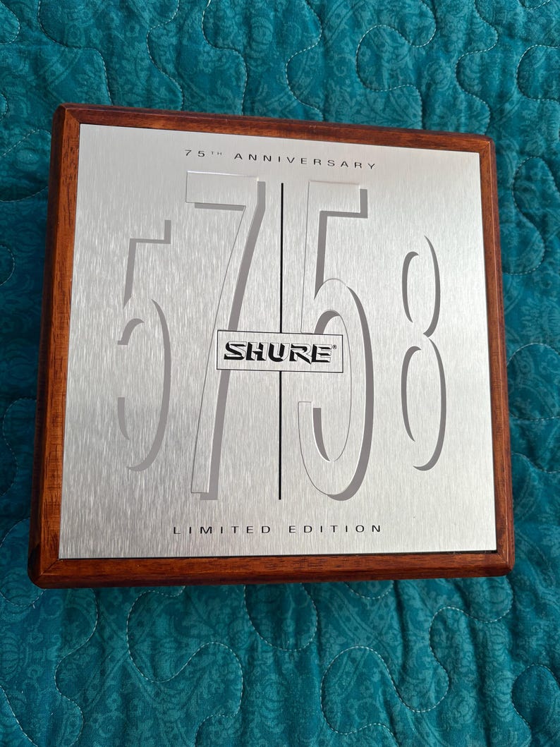 K&ouml;nnte beinhalten: Gedenktafel mit geb&uuml;rsteter silberner Oberfl&auml;che und dunklem Holzrahmen. Die Tafel zeigt die ausgeschnittene Zahl "75", das "SHURE"-Logo und den Text "75th Anniversary Limited Edition".