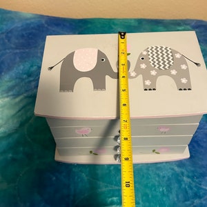 May include: A light blue wooden jewelry box with three drawers. The top of the box features a gray and white elephant design with pink accents. The drawers have a light blue background with a floral design and pink birds.