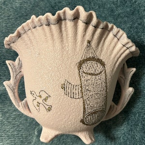 May include: A pink ceramic planter with a ruffled rim and gold accents. The planter features a bird and a cage design.