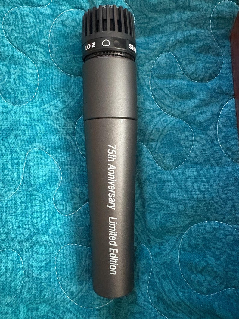 K&ouml;nnte beinhalten: Ein dunkelgraues Mikrofon mit einem schwarzen Mesh-Kopf. Auf dem Mikrofon steht der Text "75th Anniversary Limited Edition". Das Mikrofon befindet sich auf einer gemusterten blauen Oberfl&auml;che.