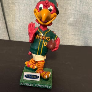 May include: A bobblehead figurine of a red bird mascot wearing a green baseball cap and a green jersey with the word HAWKS and the number 0 printed on the front. The mascot holds a brown football and stands on a green base labeled PRO-IMADE and HEISMAN HUMPHREY.