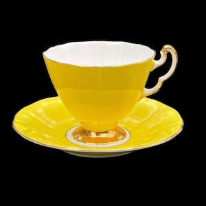 Może przedstawiać: Zestaw filiżanki i spodeczka do herbaty w kolorze żółtym z pozłacanymi zdobieniami. Filiżanka ma białe wnętrze i delikatny uchwyt.