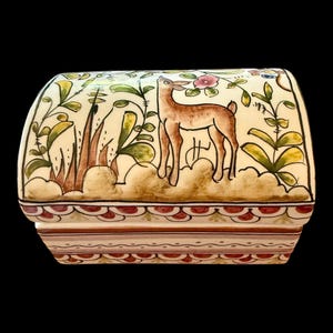 May include: A rectangular ceramic box with a curved lid featuring a hand-painted brown deer standing among green foliage and a pink flower. The base and lid edges are decorated with a repeating pattern of red scalloped shapes and brown lines.