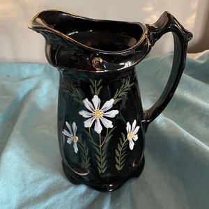 May include: Black ceramic pitcher with a gold trim and white daisy floral design.