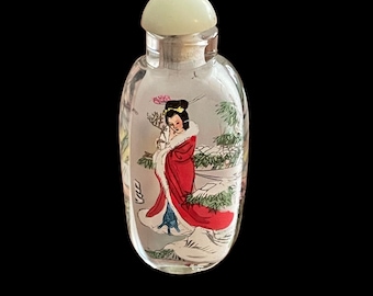May include: A small, clear glass bottle with a white lid. The bottle has a painted image of a woman in a red robe standing in a snowy landscape.