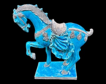 May include: A turquoise ceramic horse statue with silver accents. The horse is posed with one leg raised, and features a saddle and decorative embellishments. The statue is mounted on a square base and set against a black background.