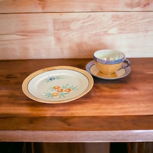 May include: A white teacup and saucer with a gold rim and floral pattern. The cup has a light blue band around the top and the saucer has a light blue rim. The teacup and saucer are on a wooden surface.