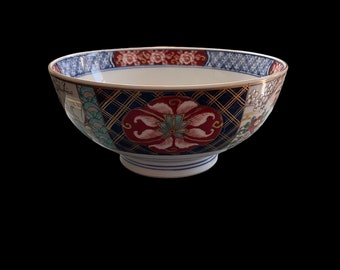 Puede incluir: Un cuenco de cerámica blanca con un diseño floral azul, rojo y dorado. El cuenco tiene un borde ancho y un fondo redondeado.