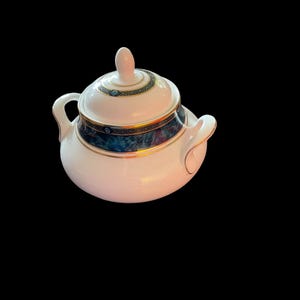 May include: A white ceramic sugar bowl with a lid. The bowl has a rounded shape with two small handles. The lid features a small knob and is decorated with a blue and gold pattern. The bowl has a gold trim.