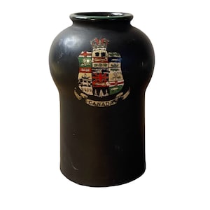 May include: A black ceramic vase with a green rim and a colorful coat of arms featuring the Canadian flag and the word "CANADA".
