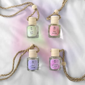 May include: Four small glass bottles with wooden caps and attached rope necklaces. Each bottle has a different colored label with text: "good days", "slow dancing", "silver lining", and "strawberry moon". The bottles are on a white surface.