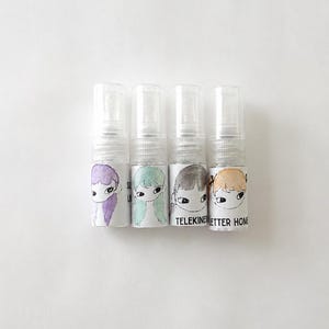 May include: Four small clear plastic spray bottles arranged in a row. Each bottle features a white label with a hand-drawn illustration of a face with different colored hair: purple, mint green, dark gray, and light orange. The text TELEKINESIS LETTER HOME is visible.