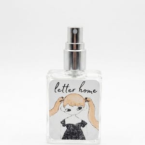 Pu&ograve; includere: Un flacone di profumo in vetro trasparente con erogatore spray argentato. L'etichetta rettangolare riporta la scritta "letter home" in corsivo nero sopra un'illustrazione disegnata a mano di una persona con lunghe codine bionde e abito nero.