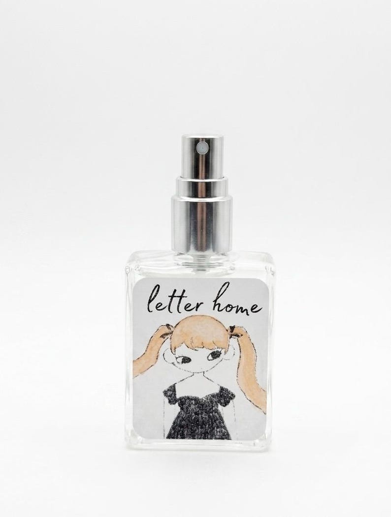 Pu&ograve; includere: Un flacone di profumo in vetro trasparente con erogatore spray argentato. L'etichetta rettangolare riporta la scritta "letter home" in corsivo nero sopra un'illustrazione disegnata a mano di una persona con lunghe codine bionde e abito nero.