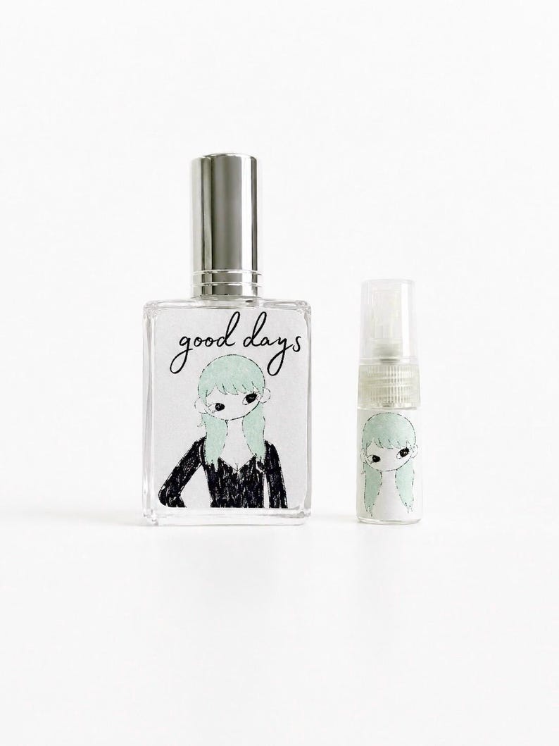 May include: A rectangular glass perfume bottle with a silver spray cap and a smaller cylindrical glass vial. Both feature a label with the text good days and a black and white illustration of a figure with light green hair wearing a black top.