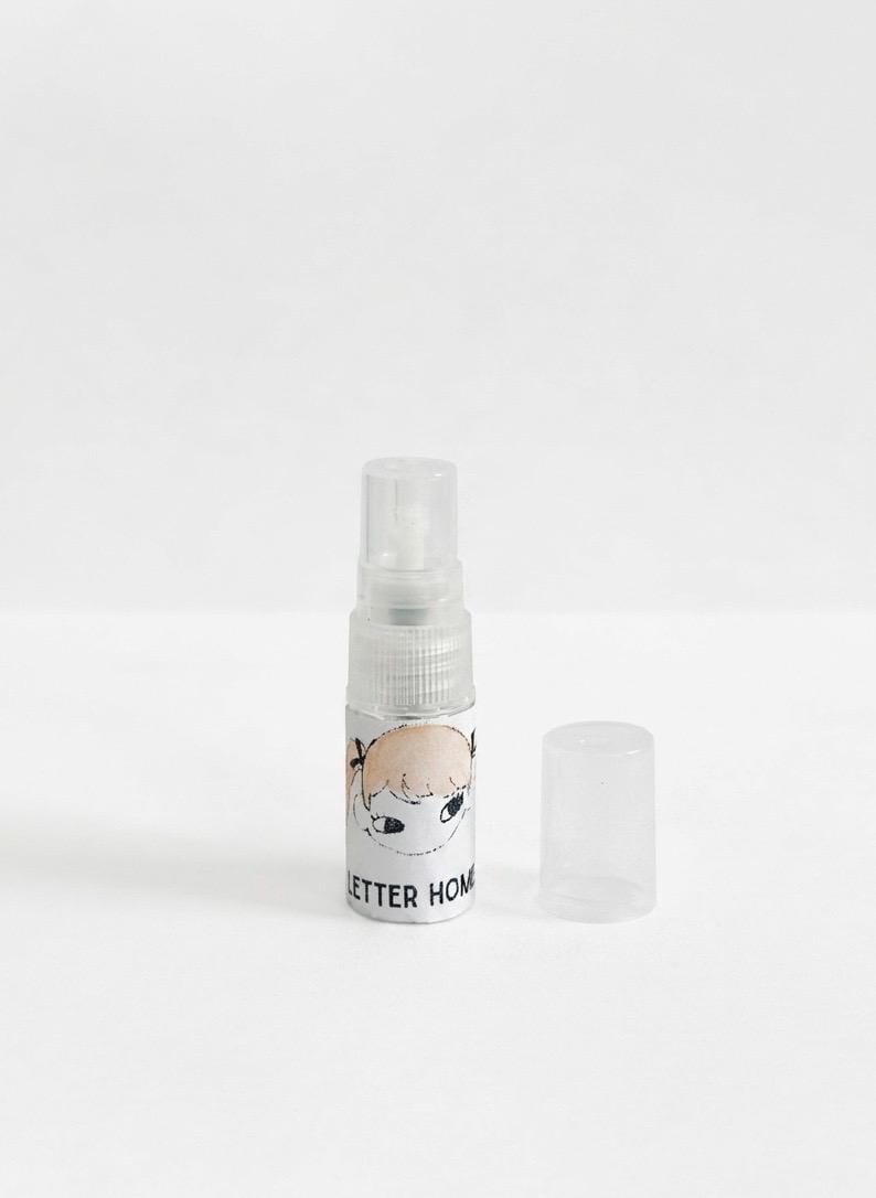 Pu&ograve; includere: Un piccolo flacone spray in plastica trasparente con tappo rimovibile. Il flacone presenta un'etichetta bianca con un'illustrazione disegnata a mano di un volto con capelli biondi e il testo nero parziale "LETTER HOM" stampato sul davanti.