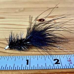 May include: A black fly fishing lure with a silver hook and black feathers. The lure is approximately 2.5 cm long and is sitting on a wooden surface next to a ruler.