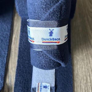 May include: Navy blue fleece wraps with a white ribbon featuring the text "Dutch Bros" and a windmill logo. The wraps are rolled up and laid out on a wooden surface. The product is designed to be used for a specific purpose.