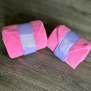 May include: Two pink fleece horse leg wraps with lavender straps. The wraps are neatly rolled and secured with a wide lavender band. The image is taken on a wooden surface.