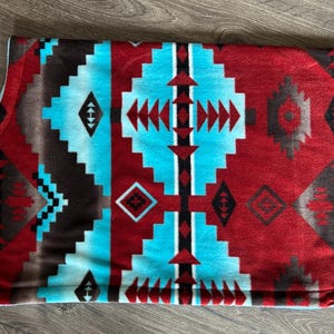 May include: A folded blanket with a Southwestern geometric pattern. The blanket features a repeating design in shades of red, turquoise, and brown. The pattern includes diamond and arrow shapes. The blanket is made of a soft, plush material.
