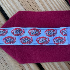 May include: A burgundy fabric wrap with a light blue ribbon featuring the Dr. Pepper logo. The wrap has a velcro closure. A roll of burgundy fabric is visible in the background. This item is likely a pet accessory.