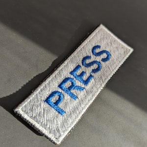 May include: A gray fabric patch with blue embroidered lettering that reads "PRESS".