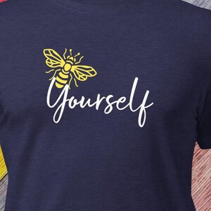 May include: Navy blue t-shirt featuring a yellow bee graphic above the word "Yourself" in a white script font. The design is simple and eye-catching, perfect for casual wear. The shirt is made of a soft, comfortable material.