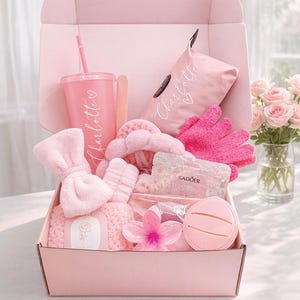 Könnte beinhalten: Eine rosa Geschenkbox gefüllt mit Pflegeprodukten. Enthält einen Becher mit Strohhalm, ein Stirnband, Handschuhe, einen Gesichtsschwamm und eine Blume. Die Artikel sind alle rosa und tragen den Namen "Charlotte".