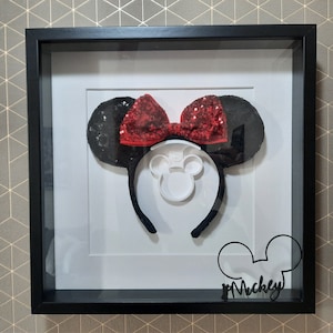 Può includere: Una scatola a ombre incorniciata in nero con un paio di orecchie di Minnie Mouse nere con un fiocco rosso di paillettes. Le orecchie sono esposte su uno sfondo bianco con una silhouette bianca della testa di Minnie Mouse. Il testo "Mickey" è scritto in nero sotto le orecchie.