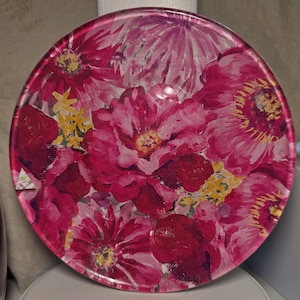 May include: A round decorative plate with a floral design. The plate features a vibrant arrangement of pink and red flowers with yellow accents. The flowers are clustered together, creating a full and colorful pattern.