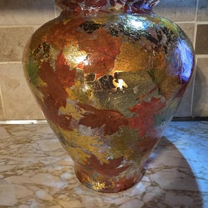 May include: A glass vase decorated with a collage of autumn leaves in shades of red, orange, yellow, and brown, interspersed with metallic gold foil accents. The vase has a wide, rounded body that tapers towards the base and a flared rim.