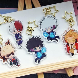 Könnte beinhalten: Eine Sammlung von Schlüsselanhängern mit Anime-Charakteren. Jeder Schlüsselanhänger zeigt einen anderen Charakter mit einzigartigen Haar- und Kostümdesigns. Die Schlüsselanhänger sind an goldenen sternförmigen Verschlüssen befestigt. Die Charaktere werden auf einer weißen Oberfläche präsentiert.