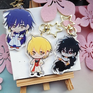 Peut inclure: Porte-clés en acrylique représentant des personnages de style anime avec des cheveux et des tenues vibrantes. Les porte-clés ont des fermoirs dorés en forme d'étoile et ronds. Les personnages sont posés sur une toile blanche sur un petit chevalet en bois, avec des décorations de fleurs roses.