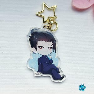 May include: A clear acrylic keychain featuring a cartoon illustration of a person with dark hair in a navy blue outfit. The keychain has a gold star-shaped clasp and a gold ring. The character is sitting with a light blue design behind them.