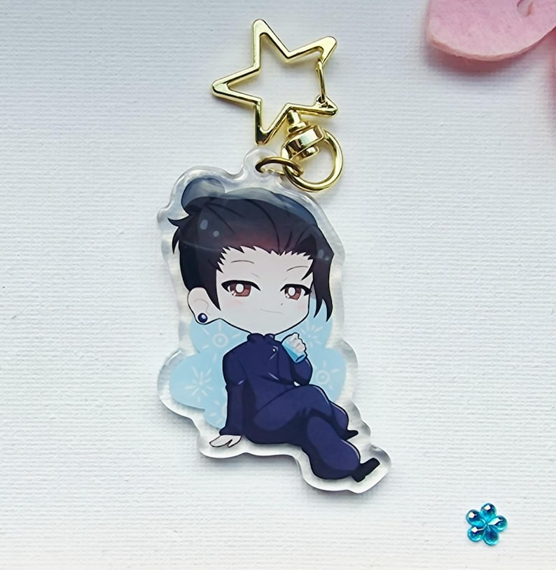 May include: A clear acrylic keychain featuring a cartoon illustration of a person with dark hair in a navy blue outfit. The keychain has a gold star-shaped clasp and a gold ring. The character is sitting with a light blue design behind them.
