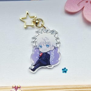 May include: A clear acrylic charm featuring a cartoon character with white hair and blue eyes, wearing a dark suit. The charm has a gold star-shaped clasp and a gold keyring. The character is sitting and holding a small red item and a drink.
